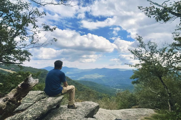 Man sitting on large rock at the hiking trail overlook looking at the long range mountain view.